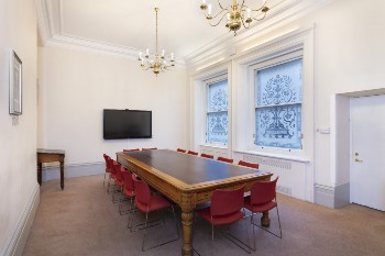 arthur holmes meeting room with white walls, a long rectangular table and red chairs and a TV screen on one wall